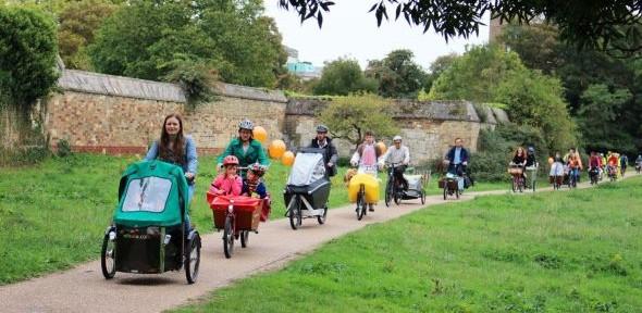 More than ten cargo bikes cycle across Coe Fen in Cambridge, carrying children holding balloons